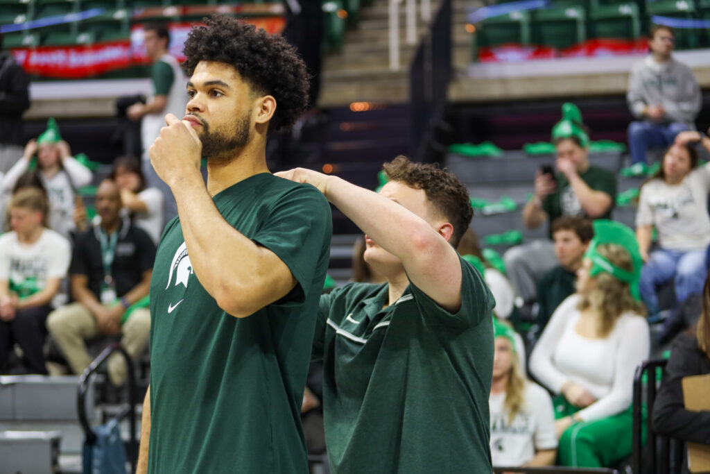 Michigan State Men's Basketball player and a coaching staff member placing Catapult device in best with the basketball court in the background.