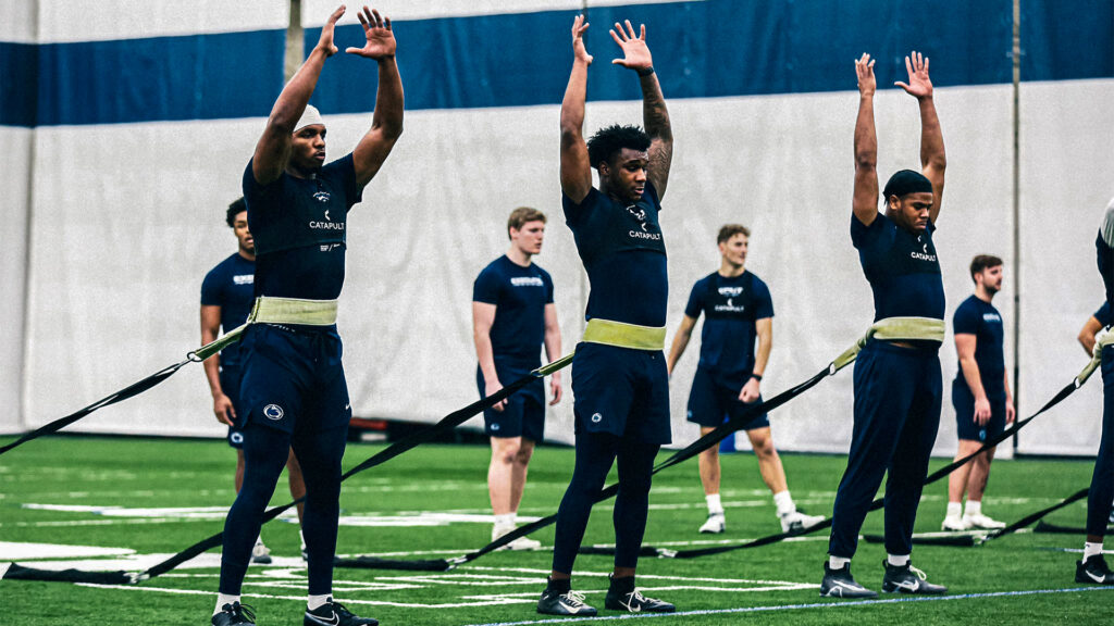 Football players in dark blue outfits performing stretching exercises with resistance bands, hands raised, in an indoor training facility.