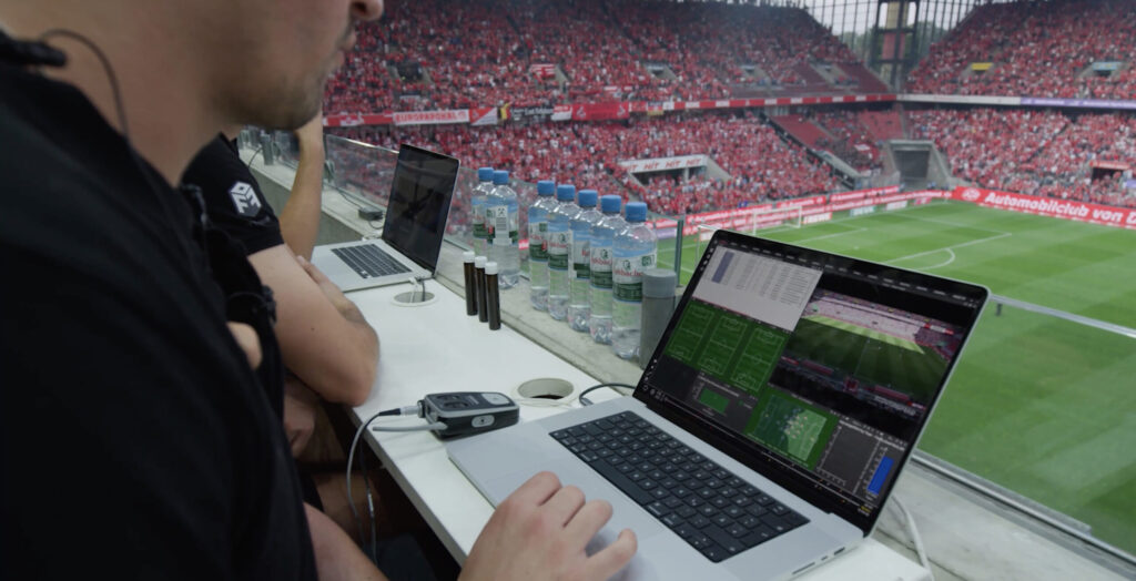 Analistas usando laptops com software de análise de desempenho durante uma partida de futebol em um estádio.