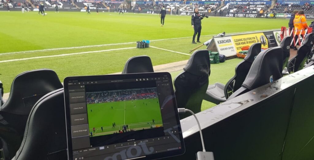 Um tablet exibindo uma partida de futebol com sobreposições analíticas, posicionado à margem de um campo de futebol.