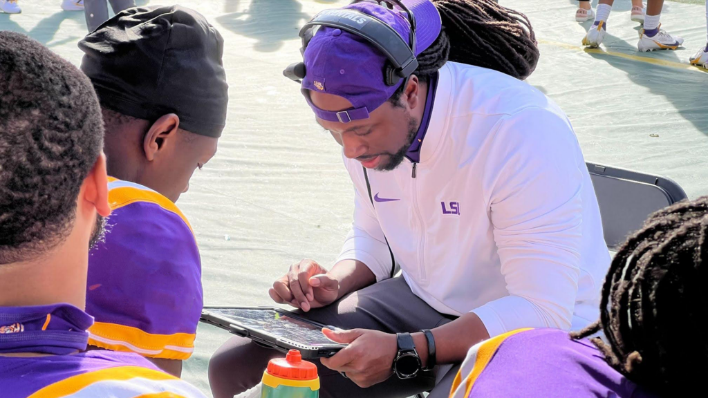 A coach wearing an LSU hat and headset reviews game footage on a tablet with a player during a football game. The players are in purple and yellow uniforms. The scene captures real-time game analysis facilitated by Catapult's sideline video solution.