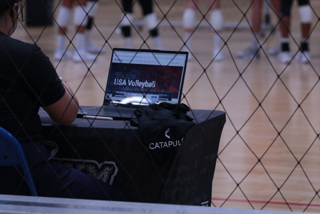 Um atleta monitorando dados em um laptop com equipamento Catapult sobre a mesa durante uma sessão de treinamento de vôlei.