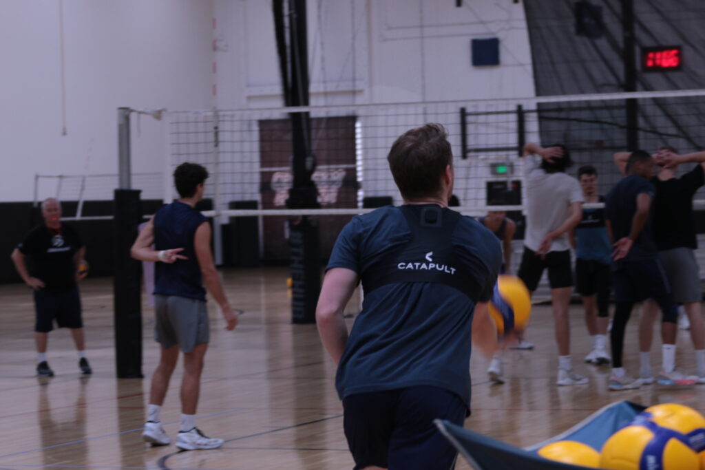 Jogadores de vôlei em uma academia, com um jogador posicionando a bola e outros usando coletes Catapulta.