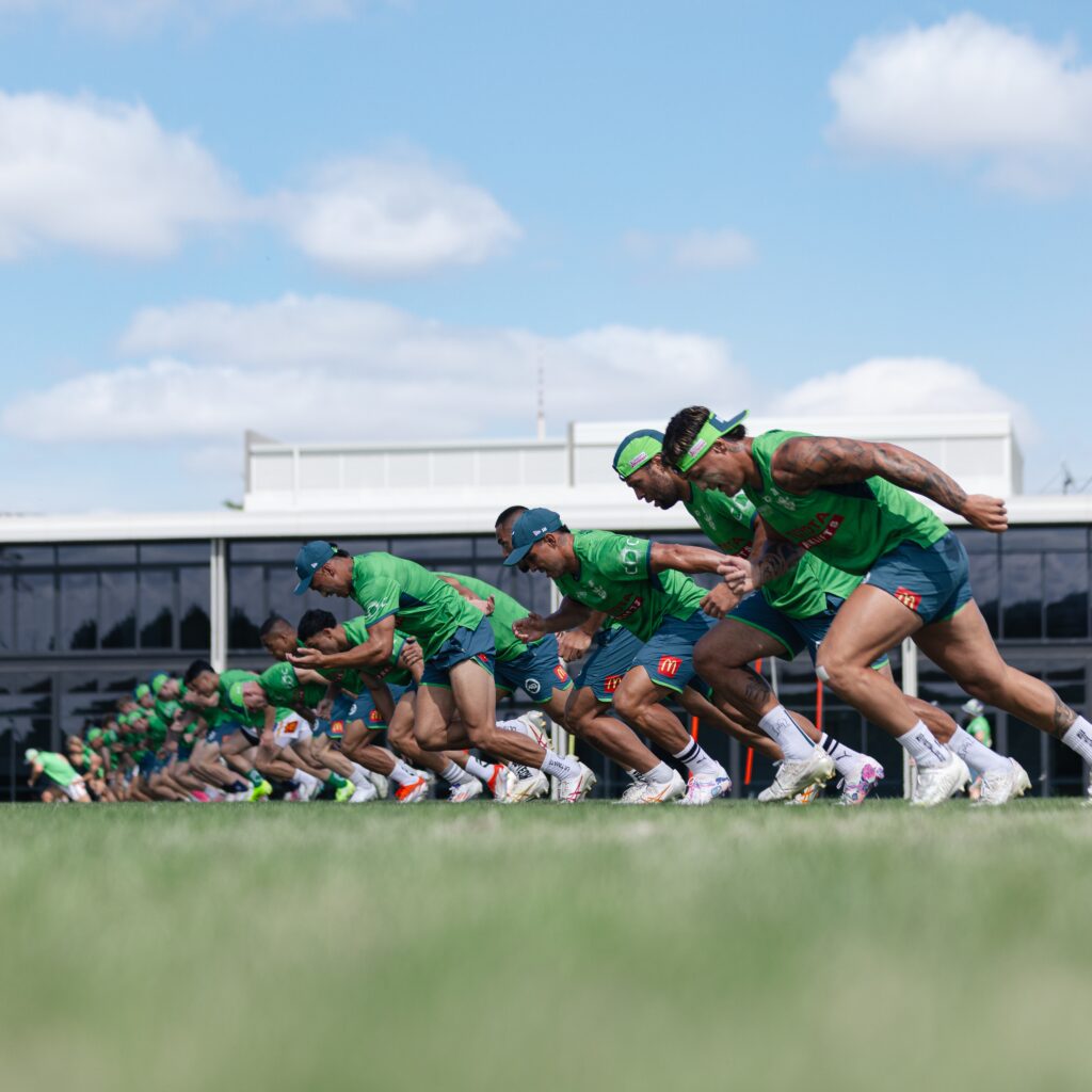 A group of Canberra Raiders players in green jerseys sprinting during a training session on a sunny day.