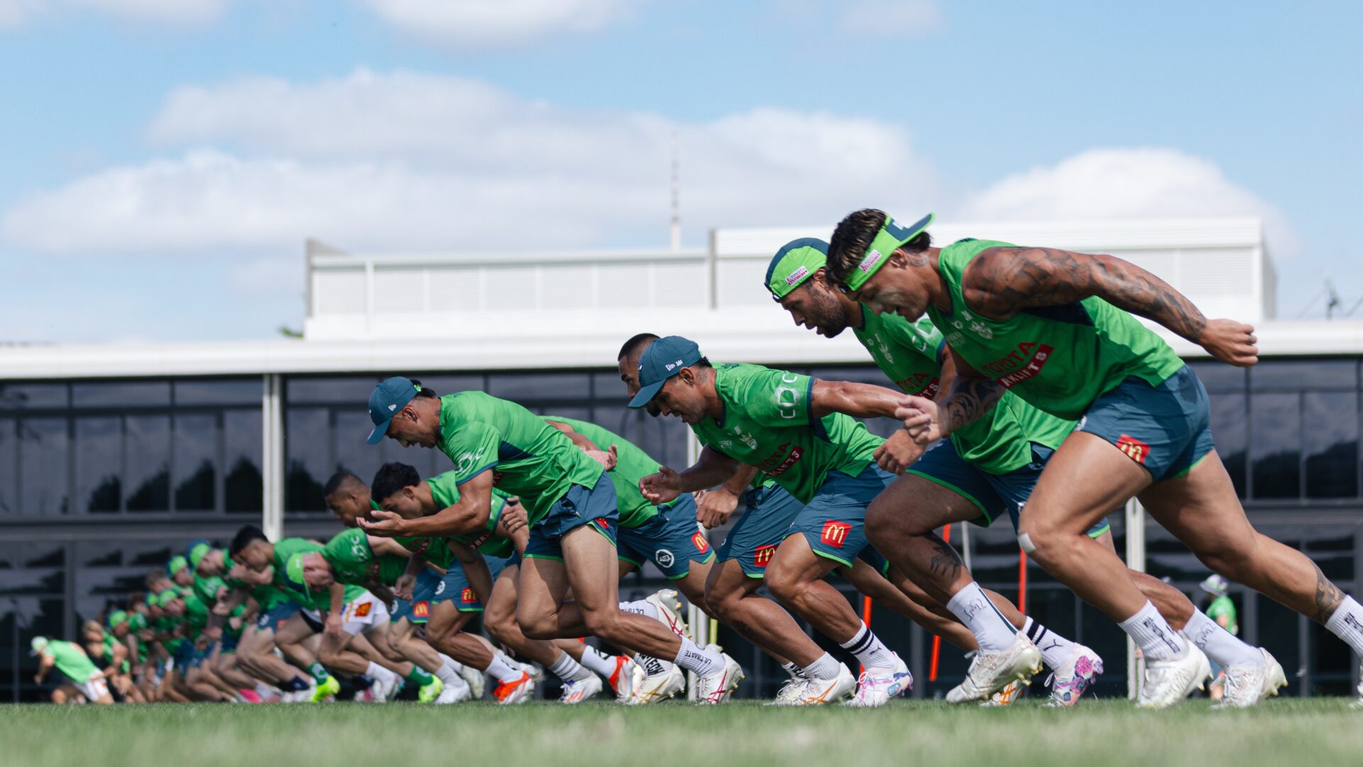 A group of Canberra Raiders players in green jerseys sprinting during a training session on a sunny day.