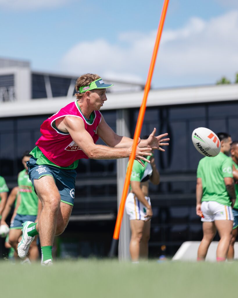 A Canberra Raiders player in a pink training vest passes a rugby ball during practice, with teammates and training equipment in the background.